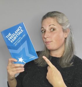 Image of Lisa Slater of LS Mentoring holding a copy of her book, The Freelance Fairytale, in her right hand and pointing to it with her left.